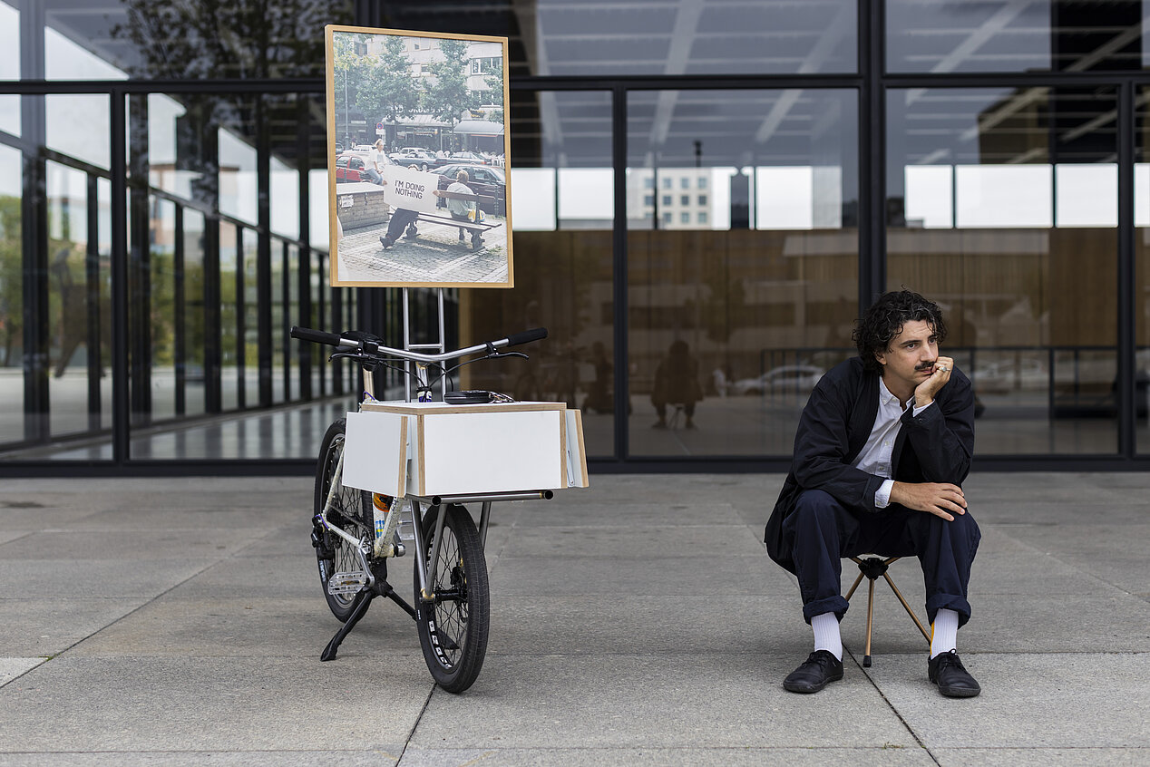 Auf dem Foto von Adrien Missika sieht man einen Mann auf einem Hocker vor einem Glasgebäude sitzen. Er hat das Kinn auf eine Hand gestützt und ist auf der rechten Seite zu sehen. Links neben ihm ist ein Fahrrad mit einem weißen Kasten. An dem Fahrrad ist ein Poster angebracht, es ist ein Foto das eine Straßenansicht zeigt. Man sieht eine Bank auf der jemand sitzt  und ein Plakat mit der Aufschrift "I'm doing nothing" hochhält.